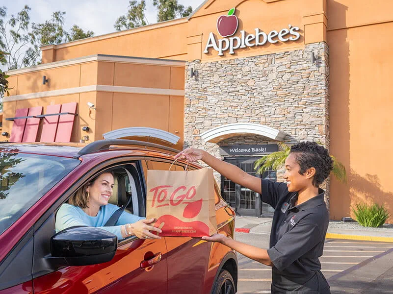 Image from the 2000s era shwoing an Applebee's female team member handing a carside-to-go order to a female in her vehicle